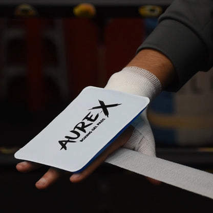 A close-up of a hand wearing a white knit glove and holding a white AUREX wrist wrap with a blue edge, set against a dark, blurred background.